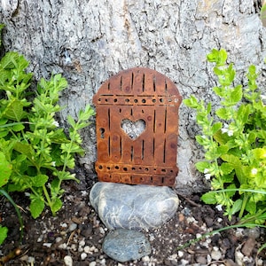 May include: A rusty metal garden decoration shaped like a birdhouse with a heart cut out in the center. The birdhouse is sitting on a rock in front of a tree trunk.