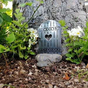 May include: A metal fairy door with a heart-shaped window, placed in front of a tree trunk, surrounded by white flowers and green foliage.