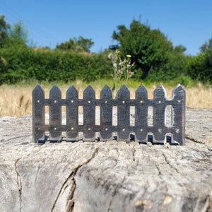 May include: A silver metal miniature picket fence with pointed tops. The fence is sitting on a weathered wooden surface.