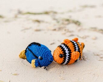 Puede incluir: Dos juguetes de peluche de peces tejidos a crochet en una playa de arena blanca. Un pez es azul con aletas amarillas y el otro es naranja con rayas blancas y negras.