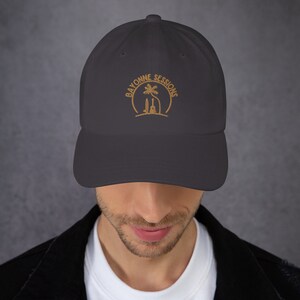 Peut inclure: Une casquette de baseball gris foncé avec un logo brodé qui dit "Bayonne Sessions" en couleur or. Le logo représente un palmier et une guitare.