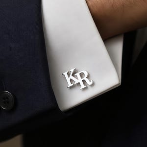May include: A silver-toned metal cufflink featuring the letters KR in a serif font, attached to the white cuff of a long-sleeved shirt. The cufflink has a polished, reflective surface and is positioned on the fabric of the shirt sleeve.