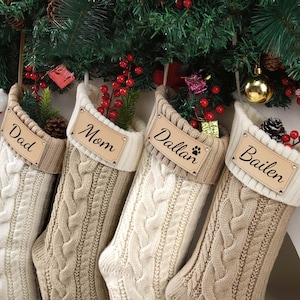 Könnte beinhalten: Vier cremefarbene und hellbraune Weihnachtsstrümpfe aus Zopfstrick hängen an einer geschmückten Tannengirlande. Jeder Strumpf hat ein personalisiertes Etikett: "Dad", "Mom", "Dallan" mit einem Pfotenabdruck und "Bailen". Festliche Weihnachtsdekoration.