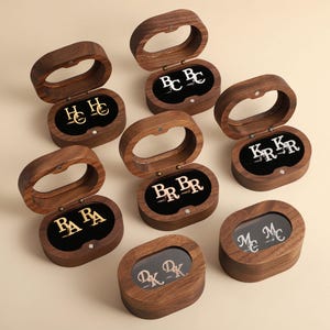 May include: Seven oval-shaped dark wood boxes with hinged lids, each containing a pair of metallic cufflinks. The cufflinks feature various monogrammed initials including HC, BC, KR, BR, RA, MC, and DK, displayed against a black velvet interior.