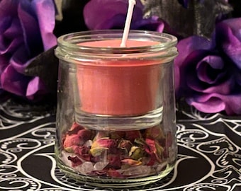 May include: A pink candle in a glass jar with dried rose petals and crystals at the bottom. The jar is sitting on a black and white patterned surface.