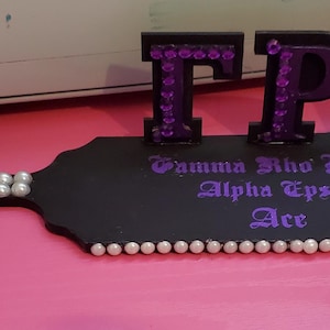 May include: A black wooden paddle with the letters TPA in purple, embellished with purple gems. The paddle has a handle adorned with white pearls and the words "Gamma Rho Lambda Alpha Epsilon Ace" in purple.