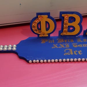 Könnte beinhalten: Ein blaues Holzpaddel mit goldfarbenen Buchstaben und Perlenakzenten. Das Paddel zeigt die griechischen Buchstaben ΦΒΛ, mit den Worten "Phi Beta Lambda", "XXI Gamma" und "Ace" in goldener Schrift. Das Paddel ist mit Perlen verziert.