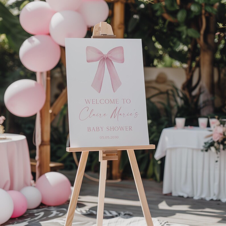 K&ouml;nnte beinhalten: Ein wei&szlig;es Schild mit dem Text "Willkommen zu Claire Marie's Baby Shower 03.05.2020" zeigt eine rosafarbene Schleife mit Aquarell-Effekt. Das Schild steht auf einem Holzstaffelei.