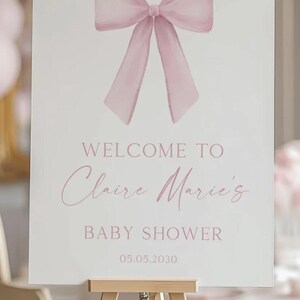 K&ouml;nnte beinhalten: Ein wei&szlig;es Willkommens-Schild mit einer rosa Schleife oben. Das Schild lautet "Willkommen zu Claire Marie's Baby Shower 05.05.2030".