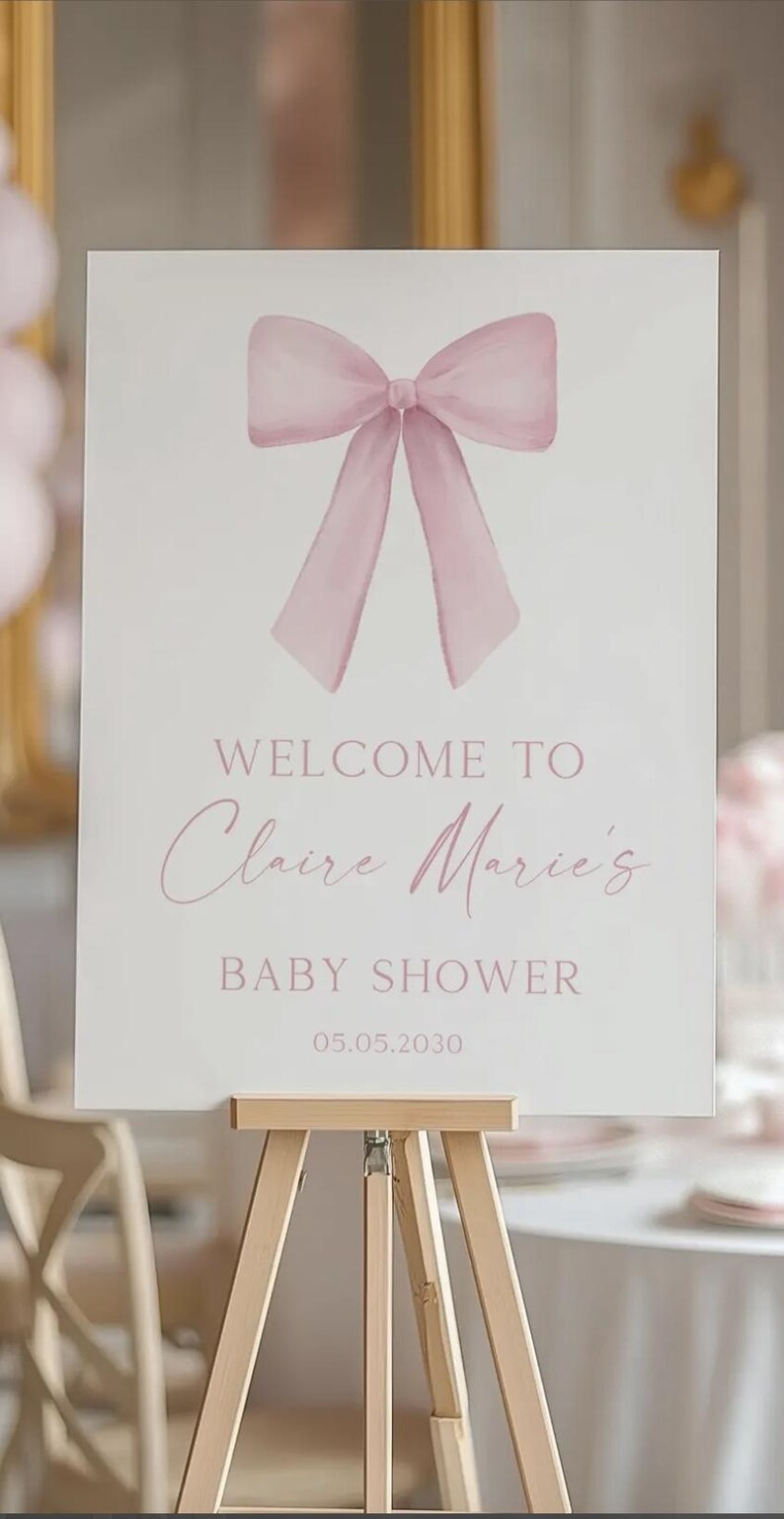 K&ouml;nnte beinhalten: Ein wei&szlig;es Willkommens-Schild mit einer rosa Schleife oben. Das Schild lautet "Willkommen zu Claire Marie's Baby Shower 05.05.2030".