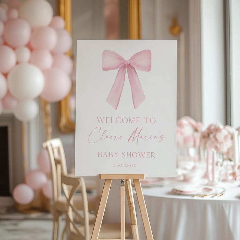 K&ouml;nnte beinhalten: Ein wei&szlig;es Willkommens-Schild mit einer rosa Aquarell-Schleife oben. Das Schild lautet "Willkommen zu Claire Marie's Baby Shower 05.05.2030".