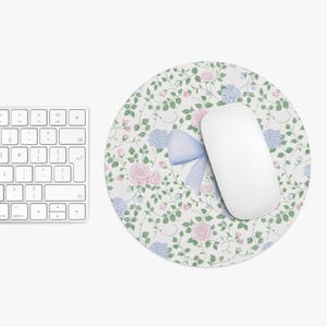 May include: A white round mouse mat with a floral pattern featuring pink and blue flowers, green leaves, and a blue bow. The mouse mat is on a white surface.