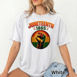 Op de afbeelding: Wit T-shirt met de tekst "Juneteenth 1865" in oranje en groen. Het shirt heeft een afbeelding van een vuist in de kleuren rood, geel en groen, in een cirkelvormig ontwerp. Het T-shirt is een casual stijl.