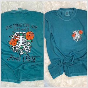 May include: Teal-colored t-shirts with a graphic design. One shirt features the text "DEAD THINGS COME ALIVE IN THE NAME OF Jesus Christ" with a rib cage design and orange roses. The other shirt has the same design on the pocket.