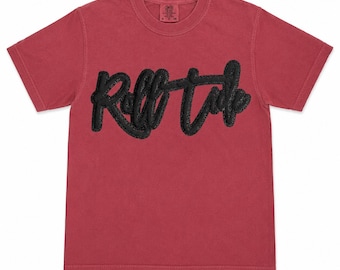 May include: A short-sleeved, crew-neck t-shirt in a muted red colour. The front features the text "Roll Tide" in a large, black, textured, raised chenille-style script font. The shirt has a standard fit with a ribbed collar and a visible brand label.