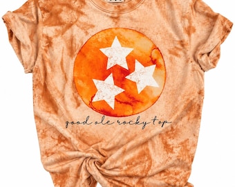 Może przedstawiać: T-shirt z krótkim rękawem i okrągłym dekoltem z pomarańczowo-beżowym wzorem tie-dye. Na środku znajduje się duże pomarańczowe koło z trzema białymi gwiazdami. Pod kołem nadrukowany jest czarną czcionką napis "good ole rocky top".