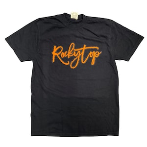 May include: A black short-sleeve t-shirt featuring the text Rocky Top printed in a bright orange, stylized cursive script across the chest. The garment has a crew neckline and a small white tag visible at the inner collar area.