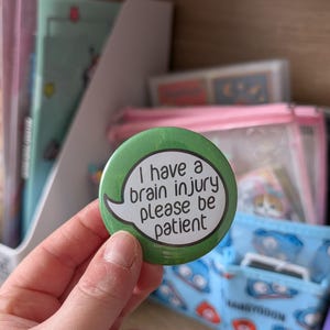 May include: A round green pin-back button featuring a white speech bubble with the black text: I have a brain injury please be patient. The button has a glossy finish and is held against a background of office supplies and storage containers.