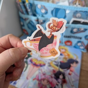 May include: A die-cut sticker featuring a cartoon bat with reddish-brown fur and large ears sitting inside a pink floral teacup. The bat has black wings and a wide, toothy grin. A tea bag tag hanging from the cup is clearly labelled with the text Bat Tea.
