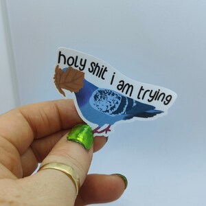 May include: A sticker depicting a blue pigeon with a brown leaf, and the text "holy shit i am trying". The sticker is held by a hand with a gold ring and green nail polish. The background is plain white.
