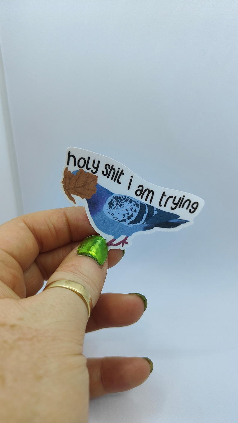 May include: A sticker depicting a blue pigeon with a brown leaf, and the text "holy shit i am trying". The sticker is held by a hand with a gold ring and green nail polish. The background is plain white.