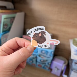 Op de afbeelding: Een sticker met een bruine en witte rat die kaas eet, met de tekst "eat cheese and sin". De sticker heeft een witte rand en een cartoonstijl.