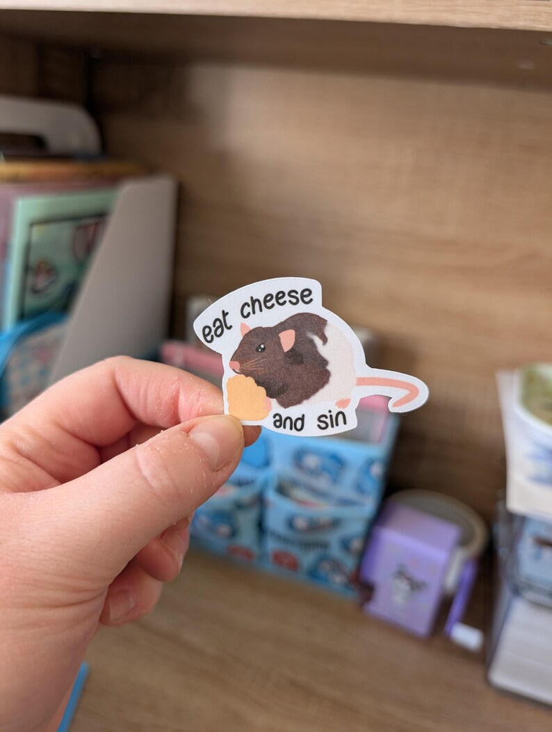 Op de afbeelding: Een sticker met een bruine en witte rat die kaas eet, met de tekst "eat cheese and sin". De sticker heeft een witte rand en een cartoonstijl.