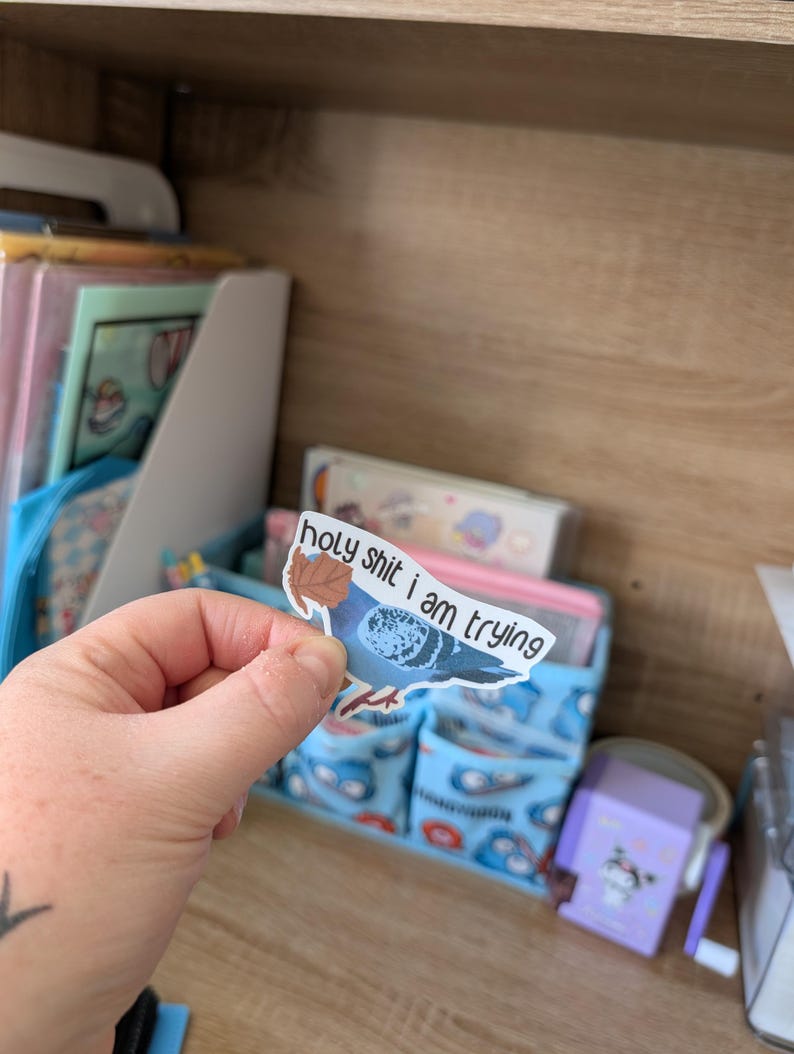 May include: A sticker showing a blue pigeon with a brown leaf and the words "holy shit i am trying". The sticker is held in a hand, with a blurred background of a desk organiser and stationery.