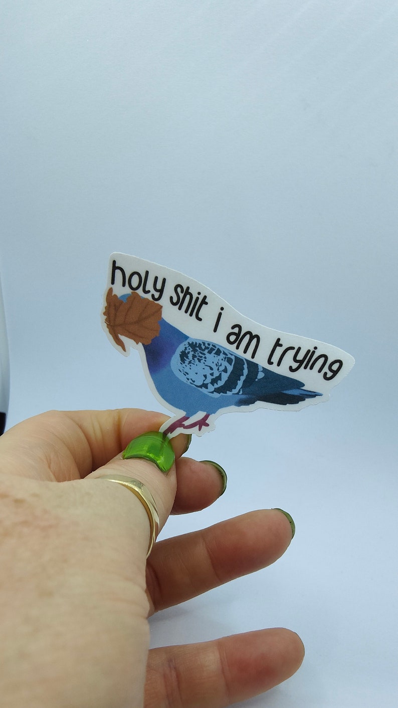 May include: A sticker depicting a blue pigeon with a brown leaf on its head. The sticker has the text "holy shit i am trying" in black. The sticker is held by a hand.