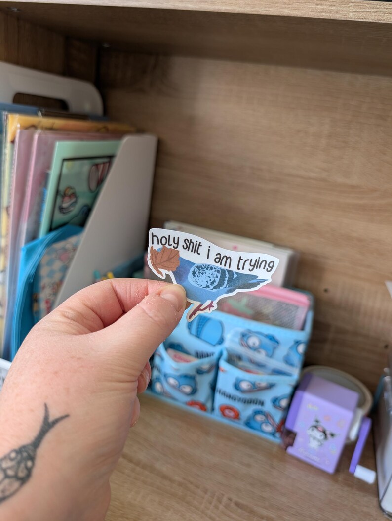 May include: A sticker depicting a blue pigeon with the phrase "holy shit i am trying". The sticker is held in a hand, with a tattoo on the wrist. A blue organiser with various items is in the background.