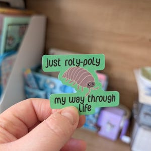 May include: A green sticker featuring the words "just roly-poly" above a brown woodlouse illustration and the words "my way through life" below. The sticker is held by a hand.