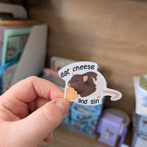 Op de afbeelding: Een sticker met een cartoonrat met bruine vacht en een stukje kaas. De tekst op de sticker luidt "eat cheese and sin". De sticker wordt in een hand gehouden.