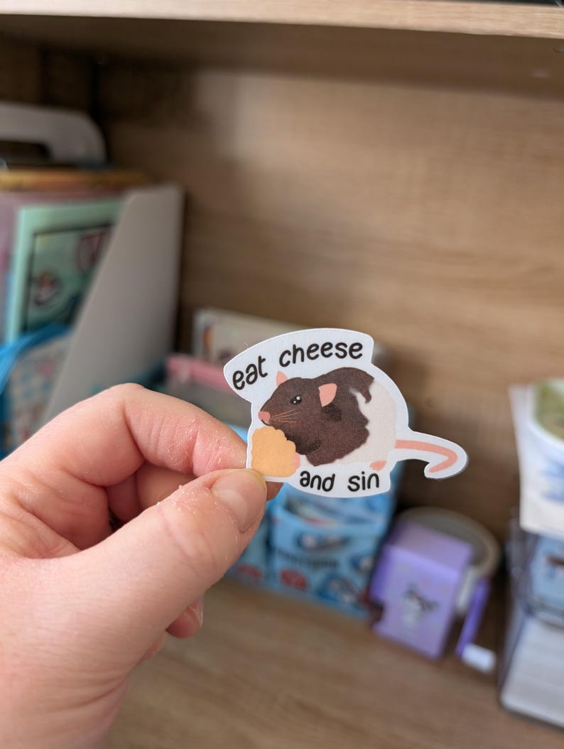 Op de afbeelding: Een sticker met een cartoonrat met bruine vacht en een stukje kaas. De tekst op de sticker luidt "eat cheese and sin". De sticker wordt in een hand gehouden.