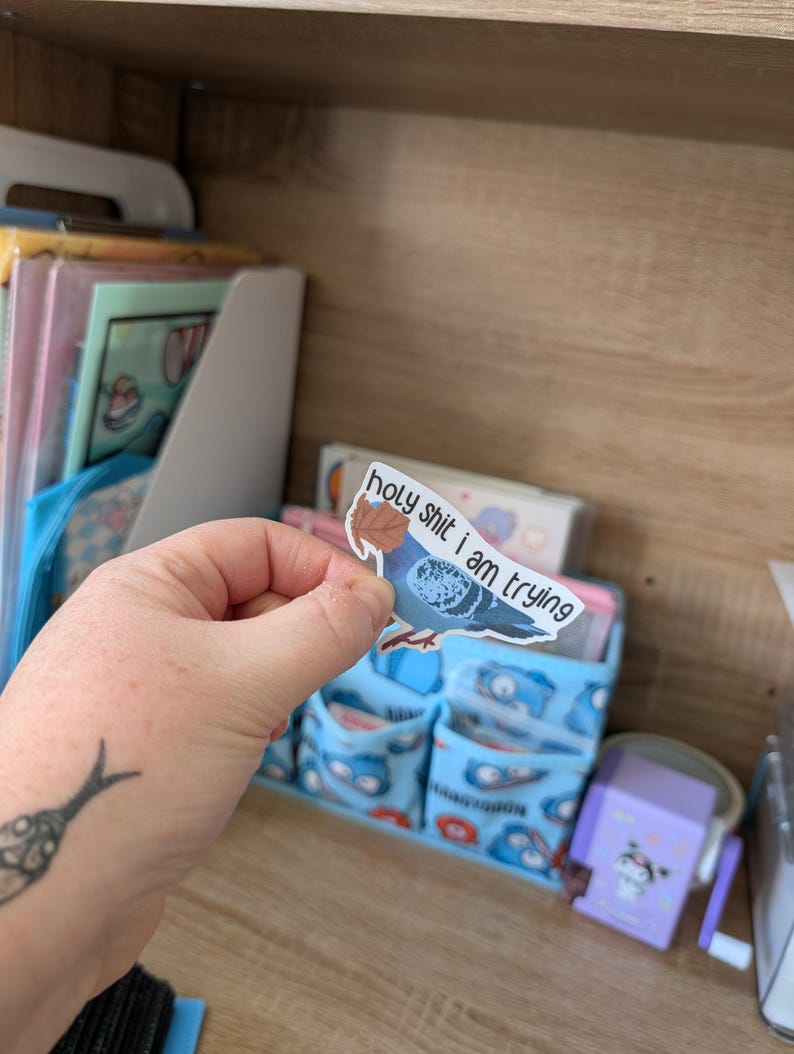 May include: A sticker depicting a blue pigeon with the words "holy shit i am trying". The sticker is held in a hand, with a tattoo visible on the wrist. The background includes a desk organiser and other stationery items.