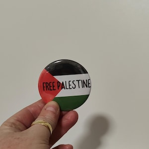 May include: A round button featuring the colours of the Palestinian flag and the text "FREE PALESTINE" in black.