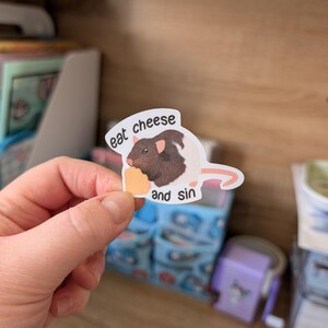 Op de afbeelding: Een sticker met een cartoonrat met bruin en wit haar, die een stuk kaas vasthoudt. De sticker heeft de tekst "eat cheese and sin". De sticker wordt door een hand vastgehouden.