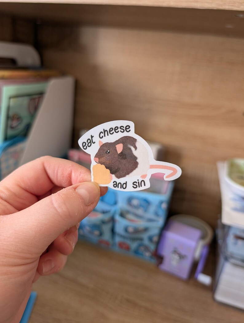 Op de afbeelding: Een sticker met een cartoonrat met bruin en wit haar, die een stuk kaas vasthoudt. De sticker heeft de tekst "eat cheese and sin". De sticker wordt door een hand vastgehouden.