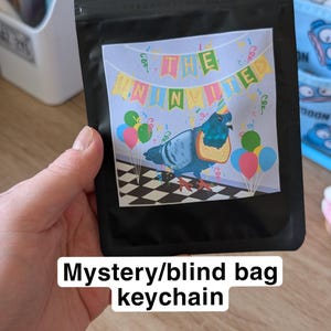May include: A black mystery blind bag containing a keychain. The label features a blue pigeon wearing a party hat and a piece of toast around its neck, with colourful balloons, confetti, and the text THE UNINVITED on a checkered floor background.