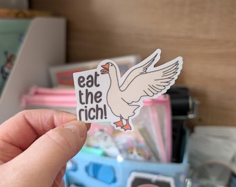 May include: A die-cut sticker featuring a white goose with orange feet and beak, depicted with wings spread wide. To the left of the goose, the text "eat the rich!" is printed in a dark brown, rounded sans-serif font against a white background.