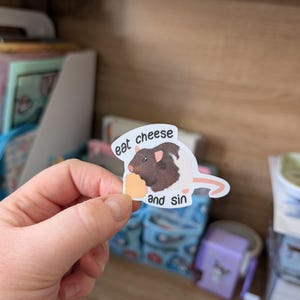 Op de afbeelding: Een sticker met een bruine en zwarte rat die een stuk kaas vasthoudt. De sticker heeft de tekst "eat cheese and sin". De sticker wordt in een hand gehouden.
