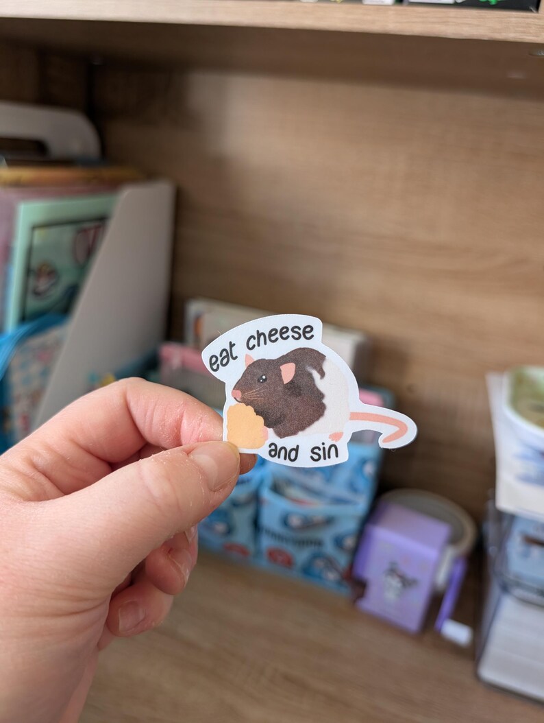 Op de afbeelding: Een sticker met een bruine en zwarte rat die een stuk kaas vasthoudt. De sticker heeft de tekst "eat cheese and sin". De sticker wordt in een hand gehouden.
