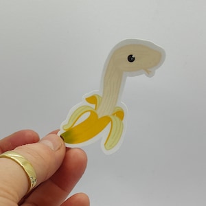 May include: A sticker of a cartoon banana with a long neck and a cute face. The banana is yellow and the neck is white.