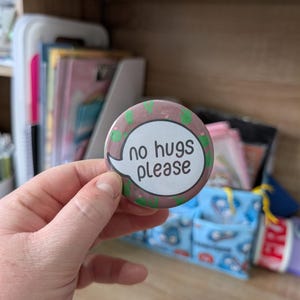 May include: A circular badge with the words "no hugs please" in a white speech bubble. The badge has a brown and green background with a decorative border. The badge is held in a hand.