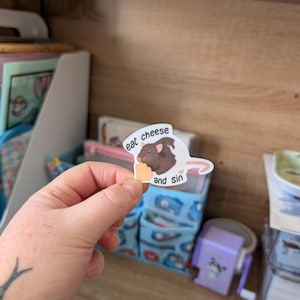 Op de afbeelding: Een witte sticker met een cartoon cavia die een stuk kaas vasthoudt. De tekst op de sticker luidt "eat cheese and sin". De sticker wordt in een hand gehouden tegen een achtergrond van een houten plank en opbergbakken.