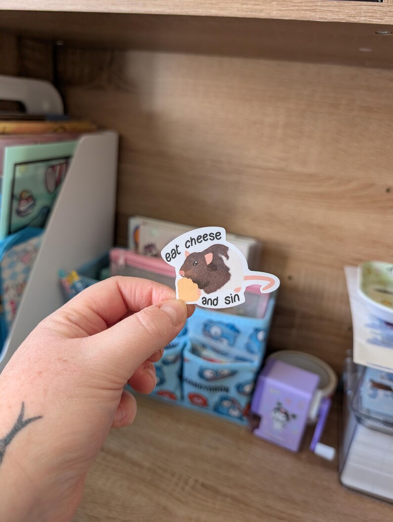 Op de afbeelding: Een witte sticker met een cartoon cavia die een stuk kaas vasthoudt. De tekst op de sticker luidt "eat cheese and sin". De sticker wordt in een hand gehouden tegen een achtergrond van een houten plank en opbergbakken.