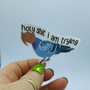 May include: A sticker depicting a blue pigeon with a brown leaf and the text "holy shit i am trying" in black. The sticker is held up by a hand with green nail polish and a gold ring.