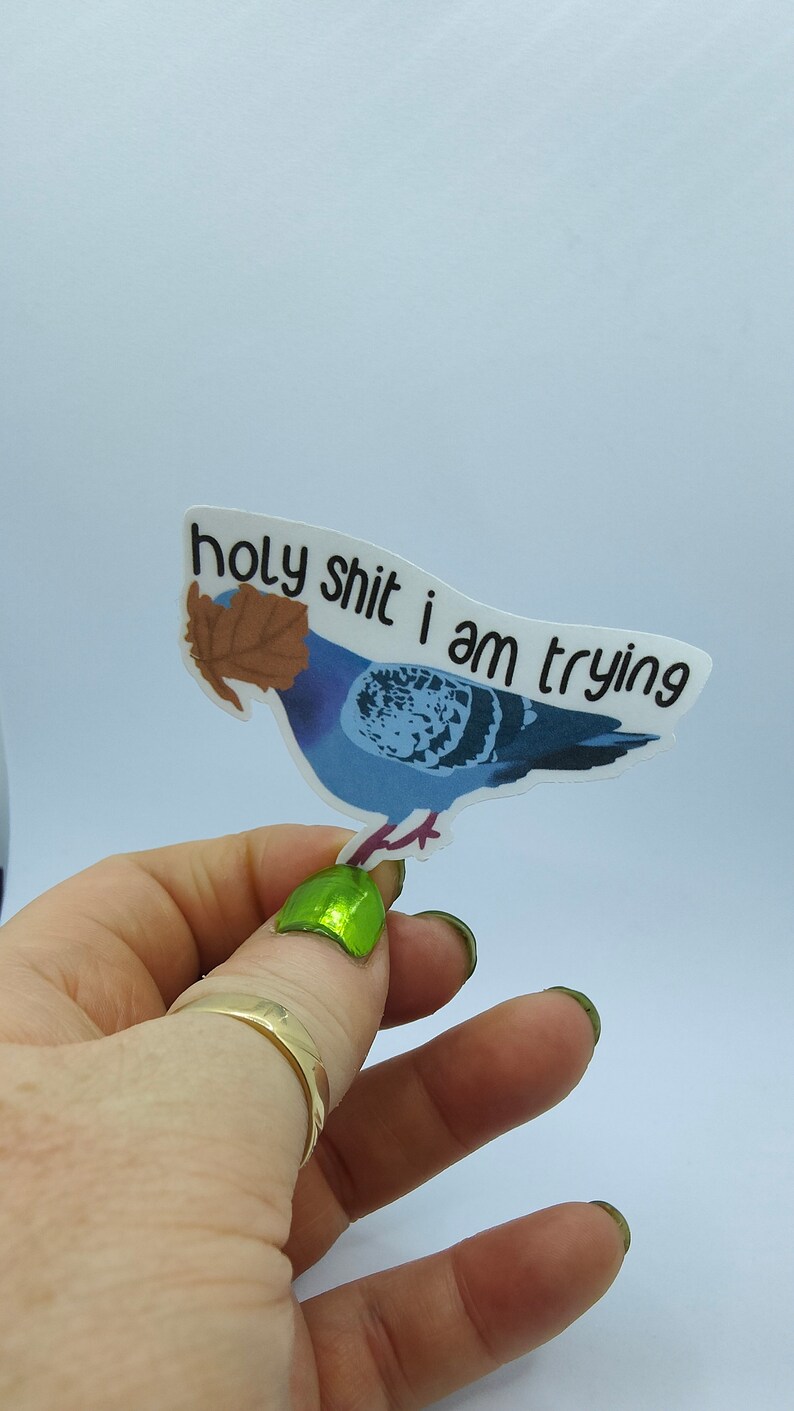 May include: A sticker depicting a blue pigeon with a brown leaf and the text "holy shit i am trying" in black. The sticker is held up by a hand with green nail polish and a gold ring.