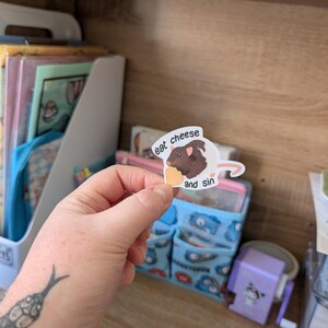 Op de afbeelding: Een sticker met een bruine cavia met de tekst "eat cheese and sin". De cavia houdt een stuk kaas vast. De sticker wordt voor een houten plank met diverse items gehouden.