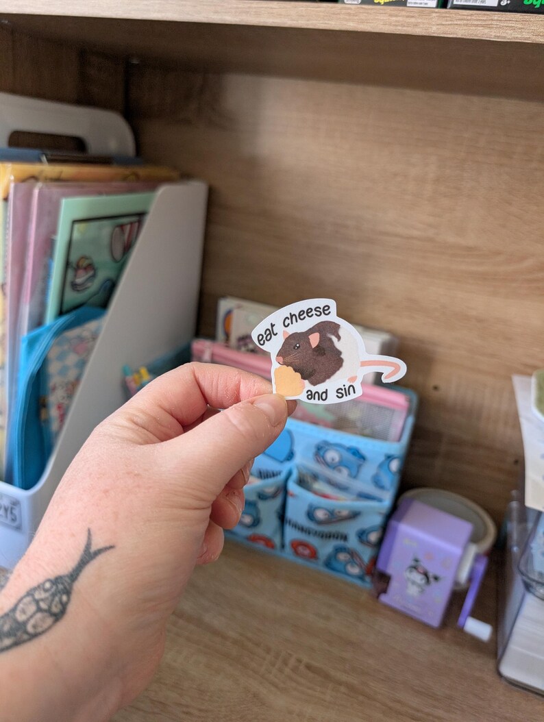 Op de afbeelding: Een sticker met een bruine cavia met de tekst "eat cheese and sin". De cavia houdt een stuk kaas vast. De sticker wordt voor een houten plank met diverse items gehouden.