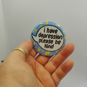 May include: A blue and yellow button with a speech bubble that says "i have depression please be kind".